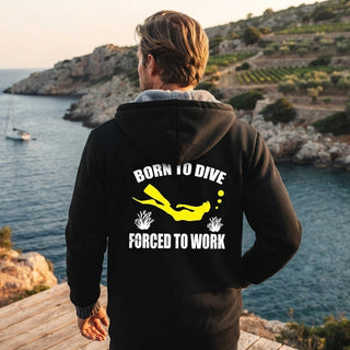 A man viewed from behind wearing a black hoodie with a yellow and white graphic on the back that reads 'BORN TO DIVE FORCED TO WORK', standing on a wooden deck overlooking a coastal sea and rocky cliffs.