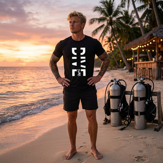 A man stands on a sandy beach at sunset wearing a black t-shirt with a large white vertical graphic and black shorts. Scuba gear is visible on a stand behind him, with palm trees and a beach bar in the background.