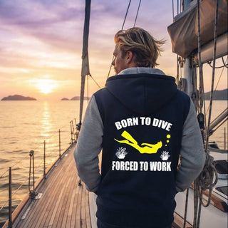 A man stands on a boat deck at sunset facing the ocean, wearing a navy and gray hoodie with a yellow scuba diving graphic and the text 'BORN TO DIVE FORCED TO WORK' printed on the back.