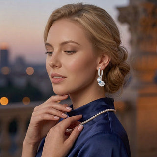 A woman with blonde hair styled in an updo wears a silver dolphin-shaped earring with light blue inlay, paired with a pearl necklace and a dark blue satin top. The background is a softly blurred outdoor cityscape at dusk.