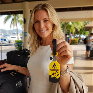 A smiling blonde woman in a white dress holds up a bright yellow and black luggage tag featuring a surfer graphic and the text 'DIVING SPECIALS'. She is standing in a covered outdoor terminal area with a black suitcase on one shoulder and a beige jacket draped off her arm.