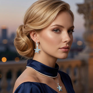 A blonde woman in a navy blue dress wearing matching starfish-shaped silver earrings and a necklace with a pearl collar, set against a softly blurred cityscape at dusk.
