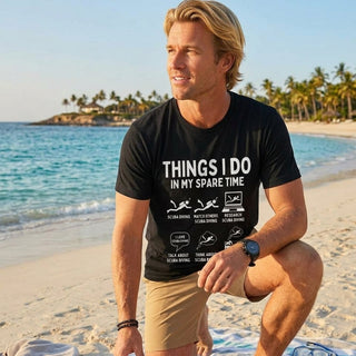 A man kneeling on a sandy beach wearing a black graphic t-shirt that reads 'THINGS I DO IN MY SPARE TIME' with swim and travel icons. He is wearing khaki swim trunks, with a blue and yellow finned pair and a towel nearby, and the ocean and palm trees in the background.