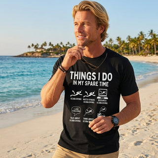A man standing on a sandy beach wearing a black graphic t-shirt with white text that reads 'THINGS I DO IN MY SPARE TIME' and tan shorts.