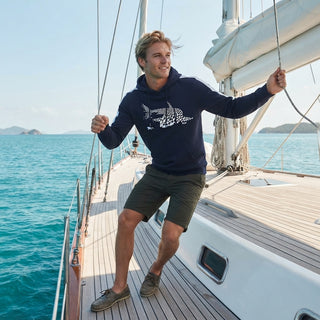 A man wearing a navy blue hoodie with a white shark graphic and dark olive shorts stands on the deck of a sailboat, holding ropes with a sunny blue sea and sky in the background.