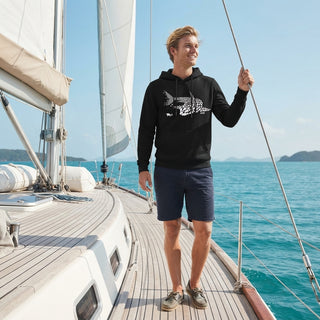 A man stands on the deck of a sailboat wearing a black hoodie with a white graphic print and navy blue shorts, looking to the side with a smile. The background features blue ocean water and a clear sky.